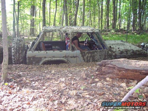 1974 Ford Bronco stuck deep in mud and leaves in a wooded area. Driver visible inside
