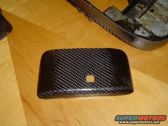 Carbon fiber trim piece with a square cutout, lying on a wooden surface