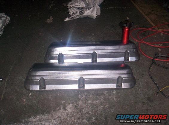 Two aluminum valve covers with a red oil cap on one, on a concrete floor