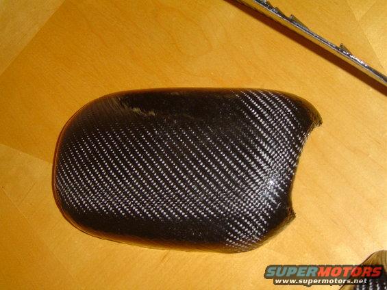 Carbon fiber side mirror cover on wooden surface. Supermotors.net logo visible
