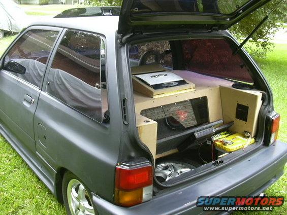 Gray hatchback car with open trunk showing custom wood-paneled sound system, subwoofers, and amplifiers