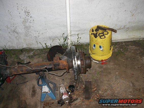 Rear axle assembly with brake rotors, supported by jack stands. A yellow bucket with a face is nearby