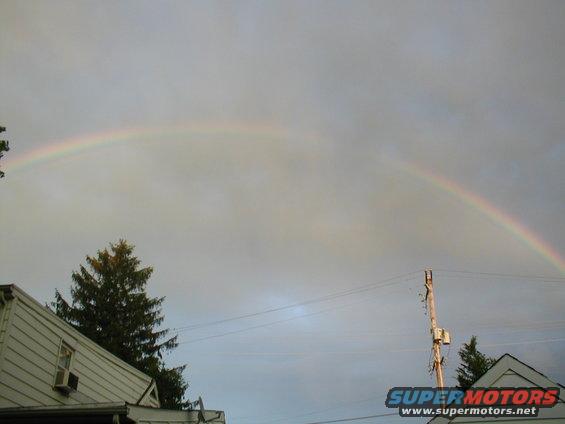 Rainbow arcs across a cloudy sky over houses and a utility pole with a Supermotors watermark