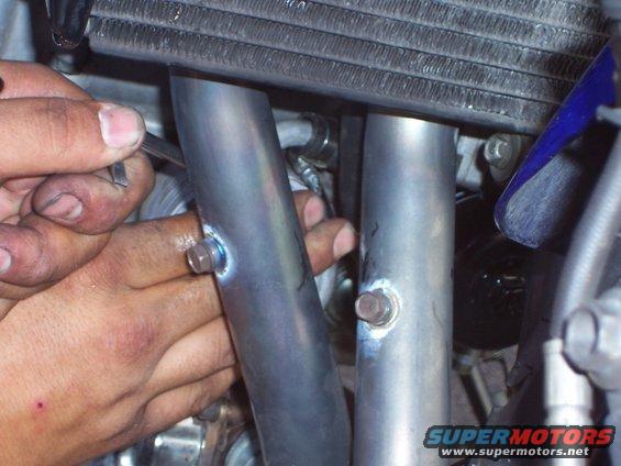 Hands working on motorcycle exhaust pipes near a radiator. Supermotors.net logo visible