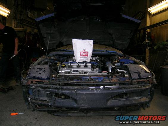 Car with hood open, engine exposed, and a Wendy's bag on top. Bumper is off
