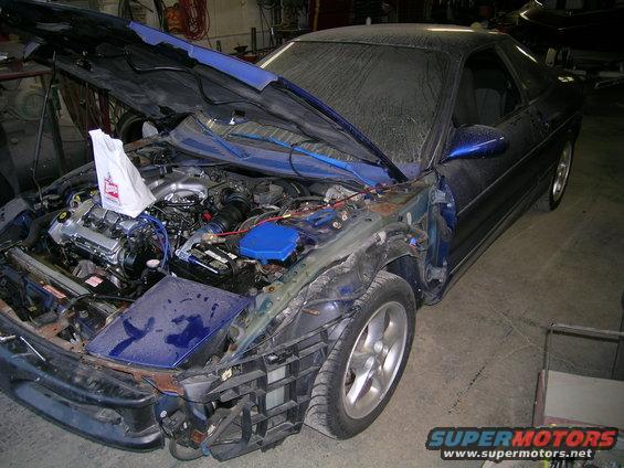 Blue coupe with hood open, showing engine and front-end damage. Dusty windshield and body. Visible Supermotors logo