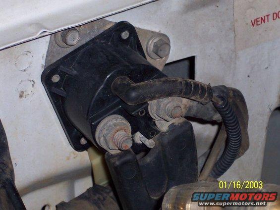 Automotive starter solenoid mounted on white vehicle bodywork, with visible rust on bolts