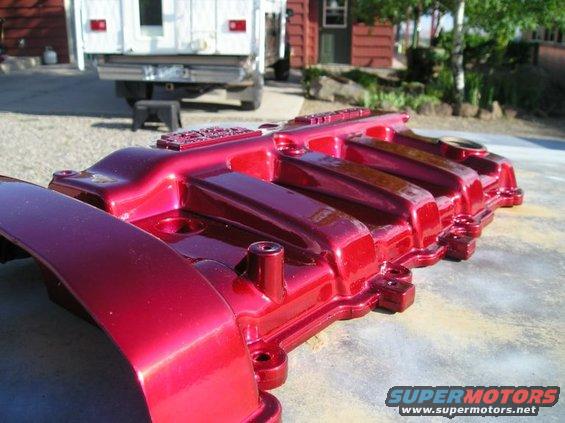 Shiny red intake manifold sitting on a concrete surface. A white truck is visible in the background