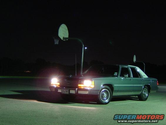 Green sedan parked at night with headlights on, basketball hoops in background