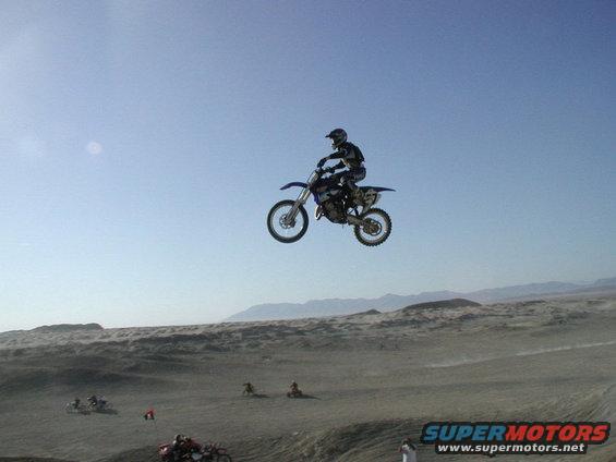 Dirt bike rider mid-air over a desert landscape. Multiple other dirt bikes visible on the ground