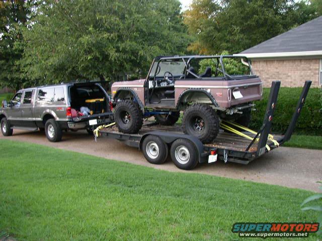 Brown Ford Bronco with roll cage on a trailer, towed by a grey Ford Excursion
