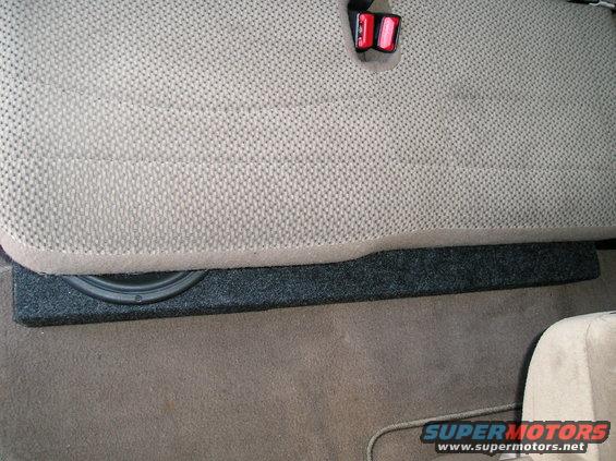 Subwoofer box under tan seat with visible speaker and seatbelt buckle