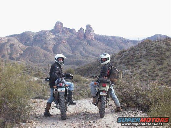 Two dual-sport motorcycles parked on a dirt road with riders, desert mountains in background