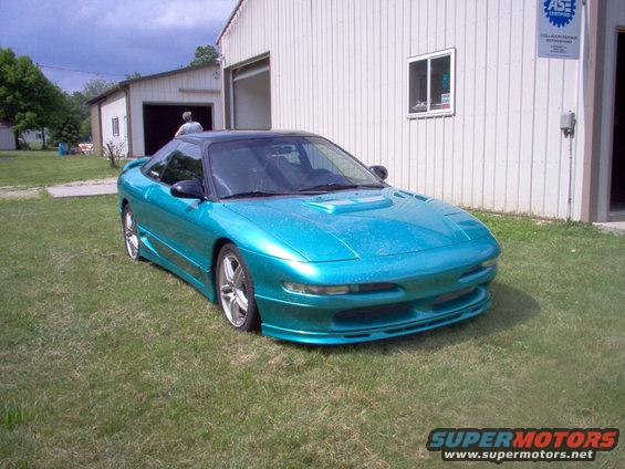 Teal coupe with black roof and aftermarket body kit parked on grass in front of a building