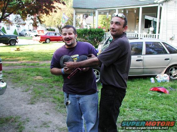 Two men in a yard, one holding what appear to be car hubcaps. A silver sedan is parked behind them