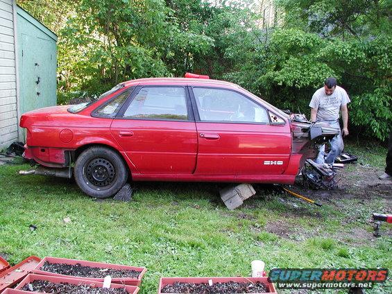 Red sedan with front end disassembled, man working on it in a grassy yard
