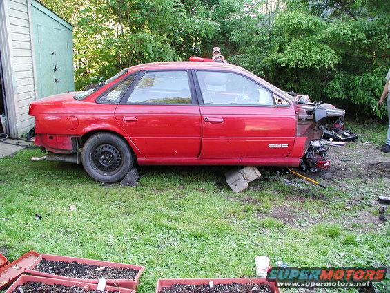 Red sedan with front end damage, missing bumper and hood, propped on blocks