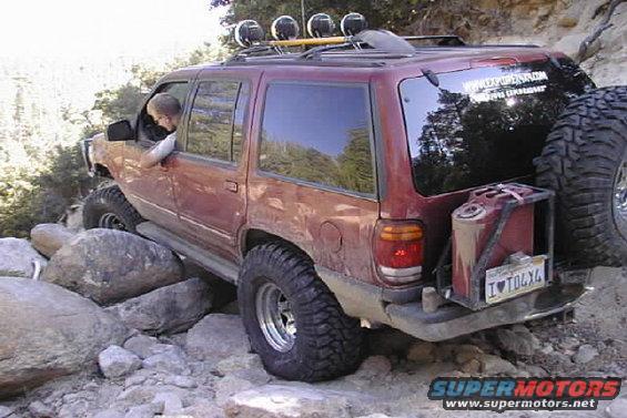 Red SUV off-roading on rocks, with spare tire and jerry can mounted on rear