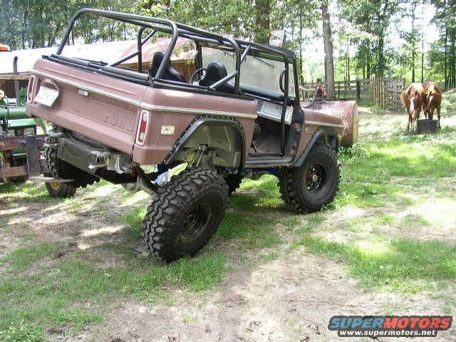 Modified first-generation Ford Bronco, brown, with roll cage and large tires, parked on grass