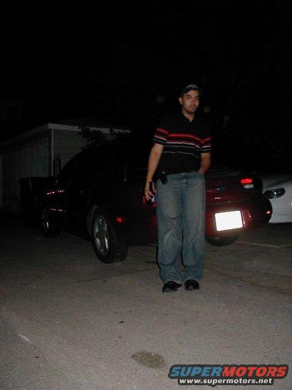 man in striped shirt and jeans stands by a red car, another white car is partially visible