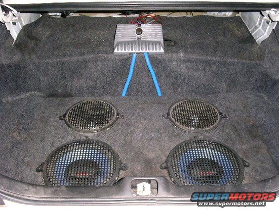 Car trunk interior with four speakers and an amplifier. Two large subwoofers are visible, along with two smaller speakers. Blue tubing connects to the amplifier