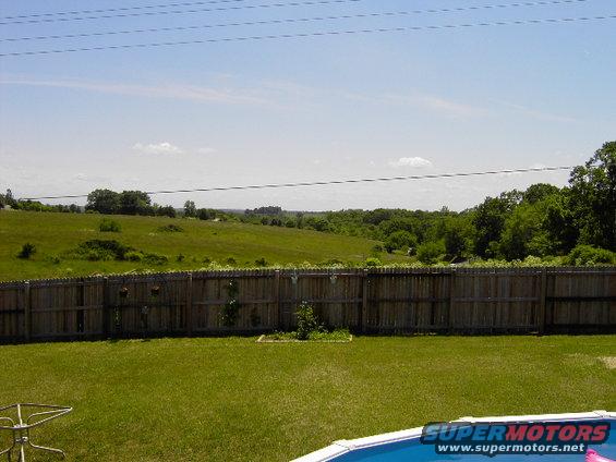 Backyard with wooden fence, green grass, and a swimming pool. Distant trees and hills under a blue sky