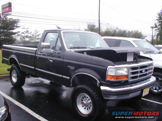 Dark blue Ford F-250 pickup truck, hood open, parked on wet asphalt. Another white SUV is parked behind it