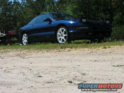 Blue 1993 Ford Probe parked on dirt with trees in background. Pop-up headlights are up