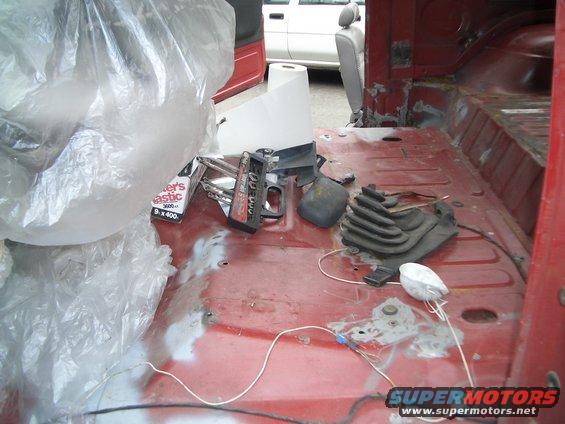Red vehicle interior floor with tools, plastic bags, and a white roll of paper. Another white vehicle is visible through the open door