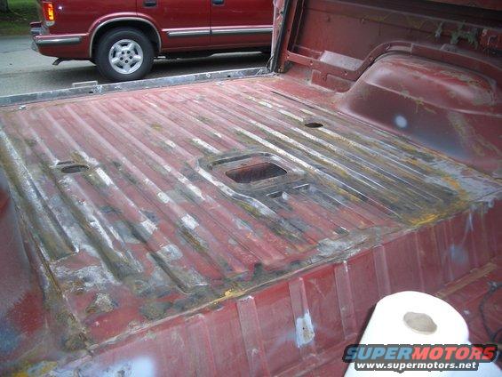 Red truck bed interior with visible rust and repair patches. A red SUV is parked behind it