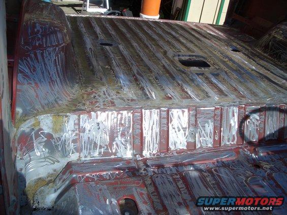 Vehicle floor pan with red primer and white scribe marks, showing ribbed texture and holes