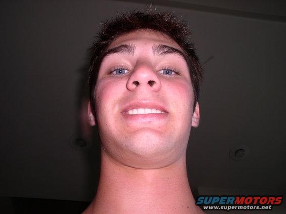 Young man looking up at the camera, smiling with teeth showing. Watermark at bottom right