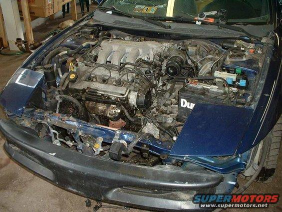 Car engine bay with hood open, showing engine, battery, and front end damage