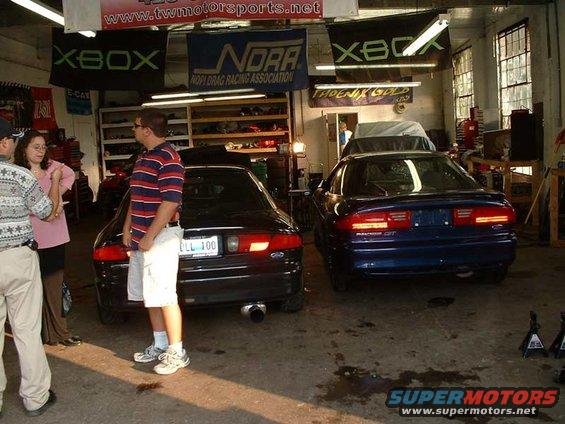 Two coupes, one black and one blue, parked in a garage. People stand near the black car