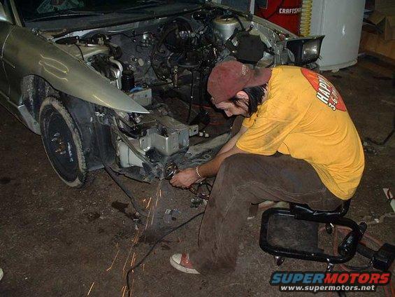 Silver car with front end disassembled, man in yellow shirt working on engine. Sparks flying