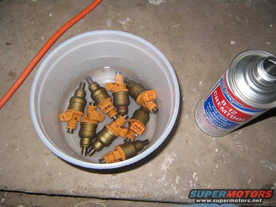 Several gold and orange fuel injectors soaking in a white plastic bowl next to a can of Berryman Chemtool