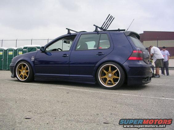 Blue hatchback car with gold rims and roof rack, parked on asphalt
