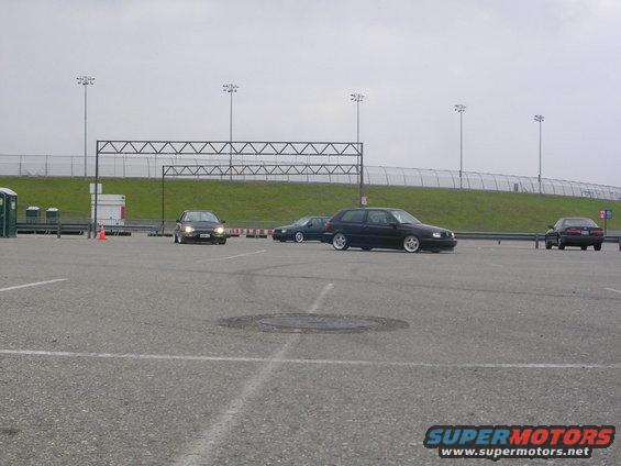 Several cars are parked and driving in a lot with overhead lights and a fence in the background