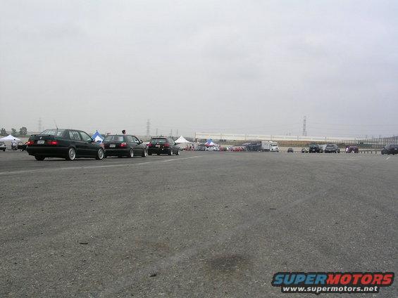 Several cars parked on asphalt under an overcast sky, with tents and trucks in the background