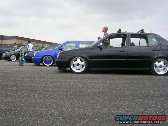 Several lowered sedans parked in a row, one black, one blue, one green. Trophies are visible in front of the green car