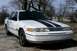 2001 Ford Crown Victoria - A white 2001 Ford Crown Victoria with black racing stripes is parked on a gravel surface.