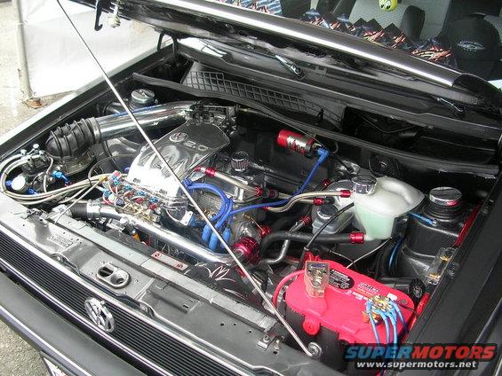 Engine bay with chrome intake manifold labeled "2.0", red battery, and blue hoses