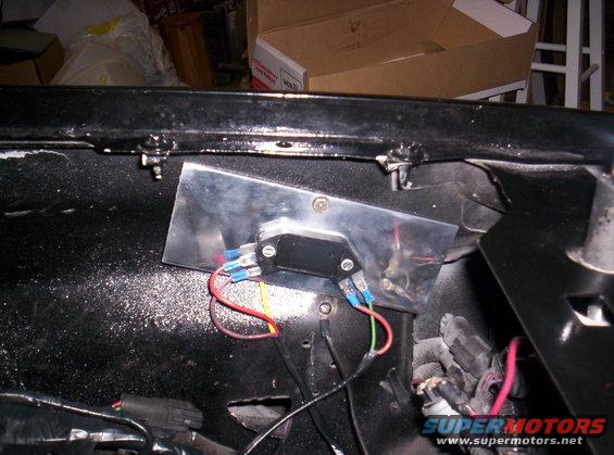 Black engine bay with a silver bracket holding a black electrical component and red, green, and blue wires