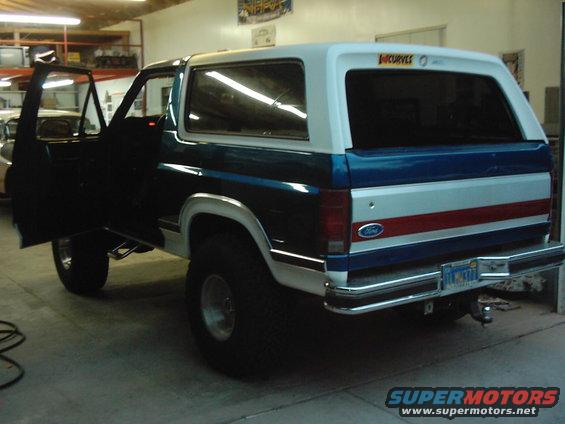 Ford Bronco with driver door open, showing side and rear. Has blue, white, and red stripes on the back