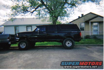 Black extended cab pickup with a camper shell, lifted with large tires, parked next to a house. Another truck is partially visible on the left