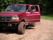 1994 Ford Bronco - A muddy red Ford Bronco sits on a dirt road surrounded by trees, with the driver's door ajar.