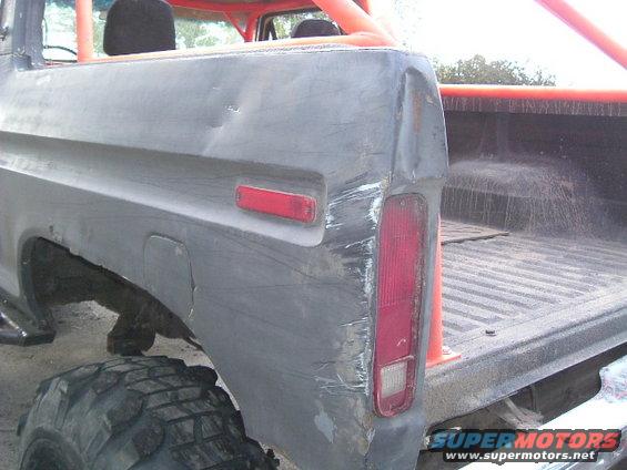 1979 Ford Bronco rear quarter panel with significant damage, dented tailgate, and missing trim. Orange roll cage visible