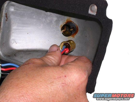 Hand with red and blue wires connecting to a brass bulb socket inside a rusty metal housing with black foam trim