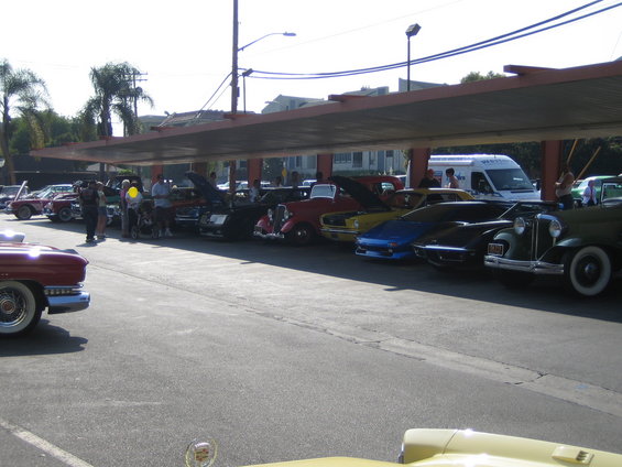 Several classic cars are parked in a row under a covered area. Some have hoods open. People are standing around them