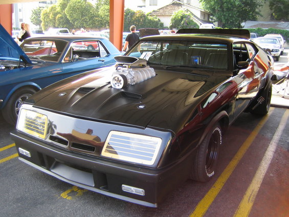 Black custom car with exposed supercharger, wide tires, and spoiler. Another blue car is visible to the left